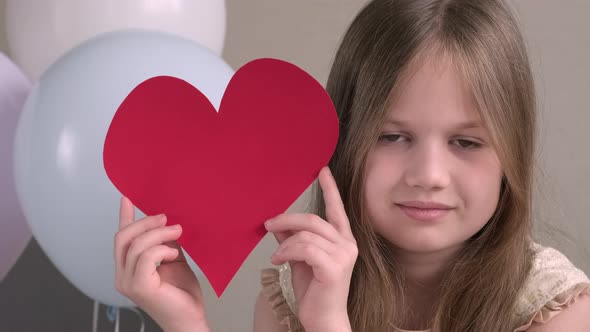 Close Up Portrait of Cute Little Girl Holding Big Red Heart on Background of Air Balloons