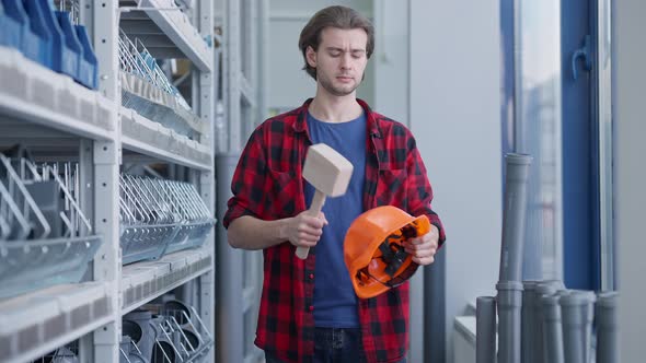 Positive Handsome Man Hitting Hard Hat with Hammer and Looking at Camera Standing in Hardware Store