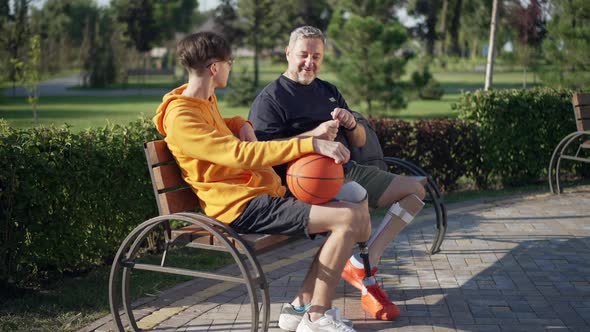 Portrait of Confident Positive Mature Father Amputee Talking with Teenage Son Sitting on Bench