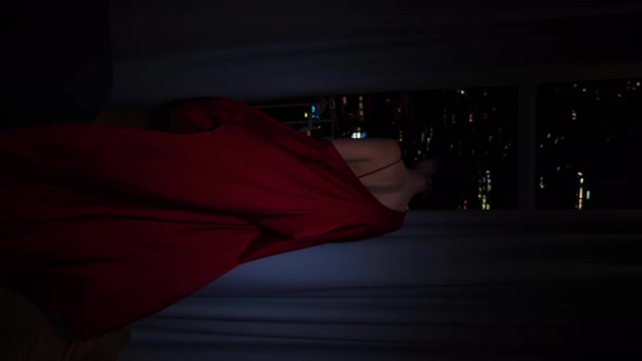 Young Woman in a Red Silk Robe Walks to the Window with Her Back the Window at the Night City with