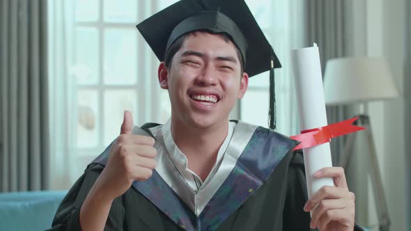 Excited Asian Man Holding A University Certificate, Smiling And Thumb Up To Camera At Home