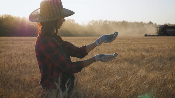 Young Woman Farmer in Hat Standing Against the Background of a Working Combine Harvester at Sunset