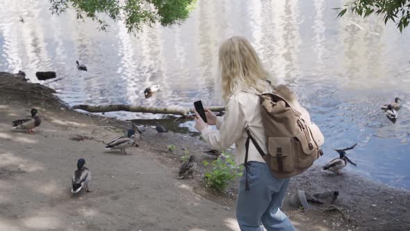 A Woman Tourist with a Large Backpack is Filming Ducks on a Pond