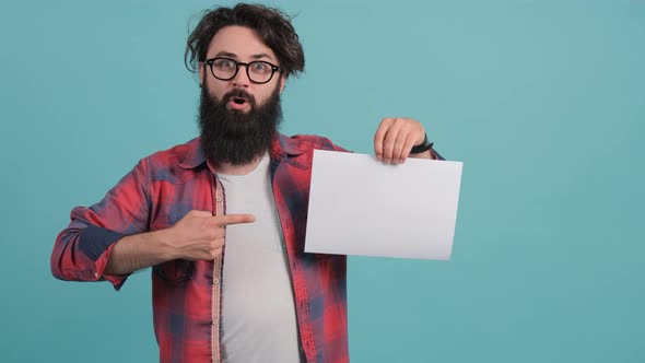 A Bearded Shocked Young Man Points To a White Sheet He Holds in His Hand.