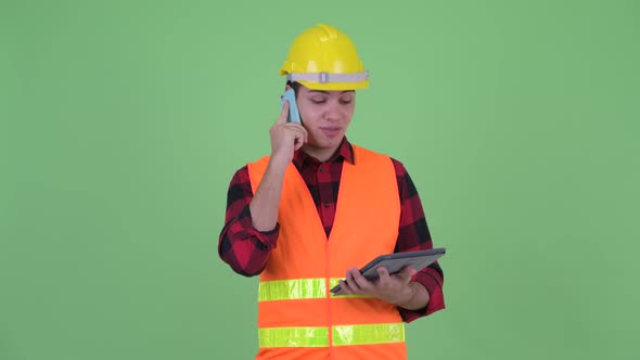 Happy Young Multi Ethnic Man Construction Worker Talking on the Phone While Using Digital Tablet