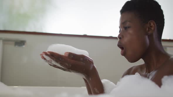 Smiling african american attractive woman taking bath and blowing out foam in bathroom