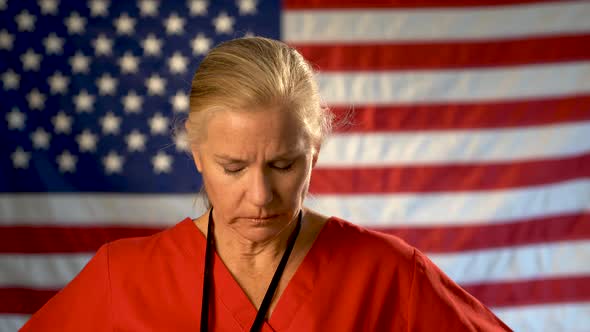 Medium tight portrait of nurse looking very worried and sad with American flag behind her.