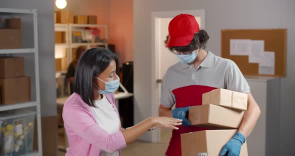 Supervisor in Face Mask Using Digital Tablet in Warehouse Talking to Courier Holding Shipping Parcel