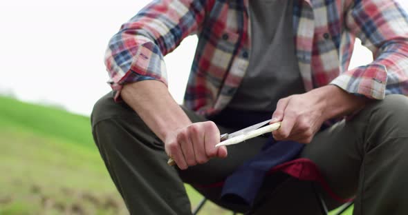 Close Up of Hands of Man Cutting with Knife a Sprig, Sitting on Camping Chair. Cloudy Sky in the