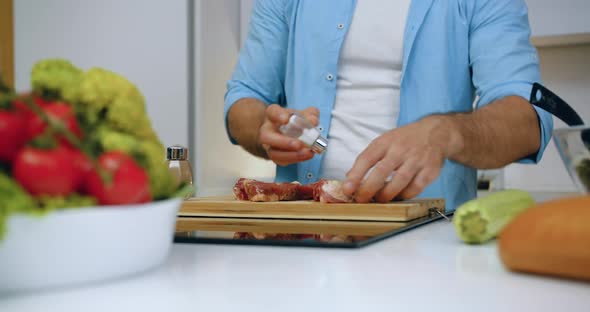 Man which Giving Salt and Pepper on Steaks on Cutting Board Before Grilling in Modern Kitchen