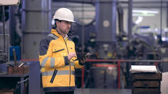 Engineer in Hard Hat is Moving Through a Heavy Industry Factory with a Tablet Computer