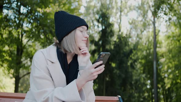 Young Girl Sitting on a Park Bench in Autumn Texting on the Phone and Smiling