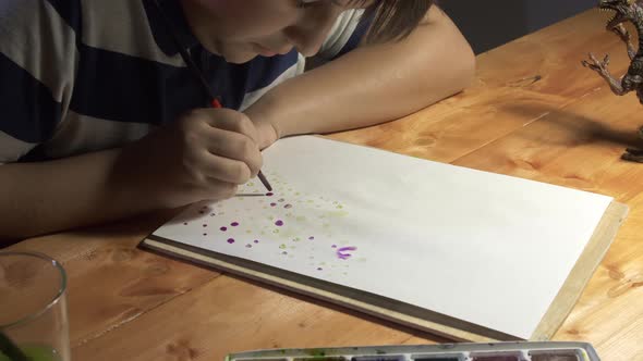 A Boy Draws Watercolor Sitting at a Table Under a Desk Lamp