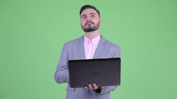 Happy Young Bearded Businessman Thinking While Using Laptop