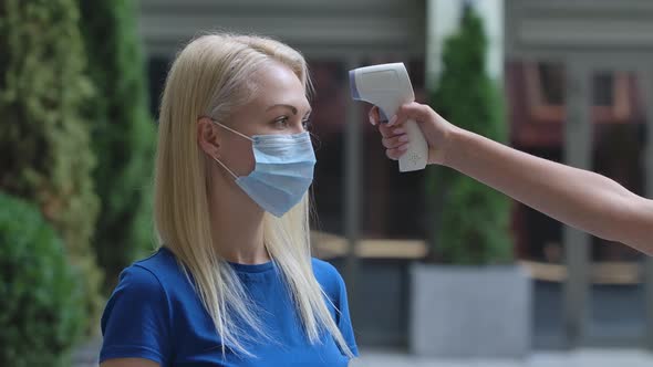 Portrait of a Charming Young Woman in a Protective Medical Mask Whose Temperature Is Measured with a