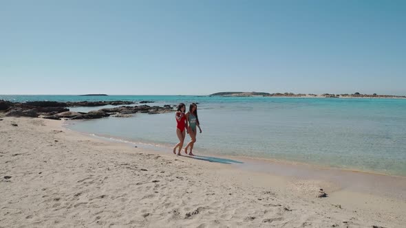 Pretty Female Friends Walking Holding Hands By Sea on Sandy Tropical Beach in Swimsuits