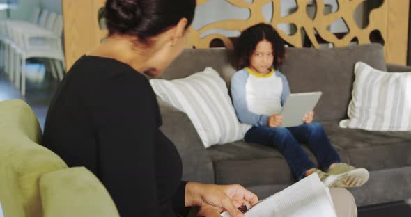 Side view of Caucasian woman reading magazine and African american boy using digital tablet on sofa