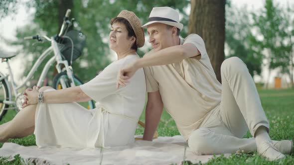 Elegant Mid-adult Caucasian Couple Sitting on Blanket in Summer Park and Chatting. Portrait of Happy