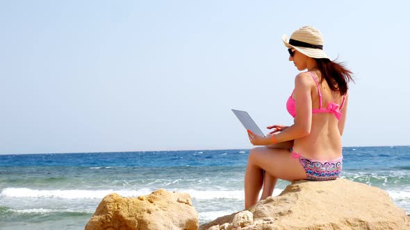 Young Woman in Swimsuit, Sun Hat and Sunglasses, Uses Laptop, Sitting on Stone on Beach By the Sea