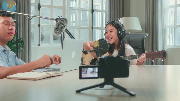 Asian Kid Girl In Headphones Playing The Guitar And Singing While Recording Podcast