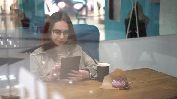 A Young Woman is Sitting Behind a Glass in a Cafe with a Tablet in Her Hands