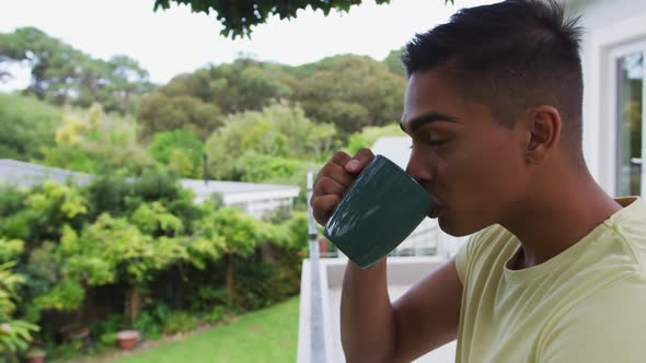 Mixed race man standing on balcony drinking a mug of coffee looking at garden