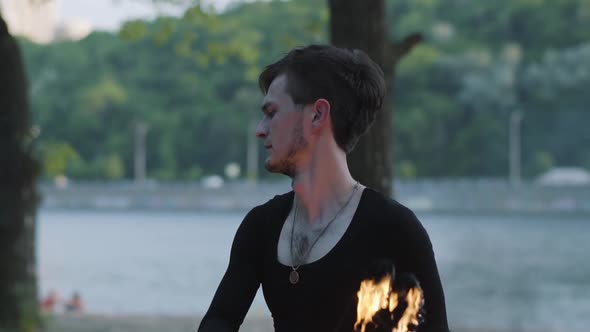 Portrait of Young Slim Man Performing a Show with Fire Fan Standing on Riverbank in Front of Trees