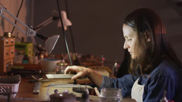 Profile of focused caucasian female jeweller sitting at desk, making jewelry in workshop