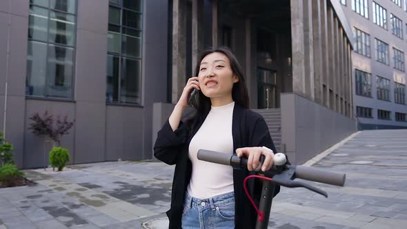 Asian Girl in Casual Clothes Enjoying Her Phone Conversation while Walking with Own Electric Scooter