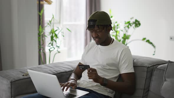 Young Black Man Holds Plastic Bank Card Sitting on a Comfortable Sofa Shopping Using a Laptop