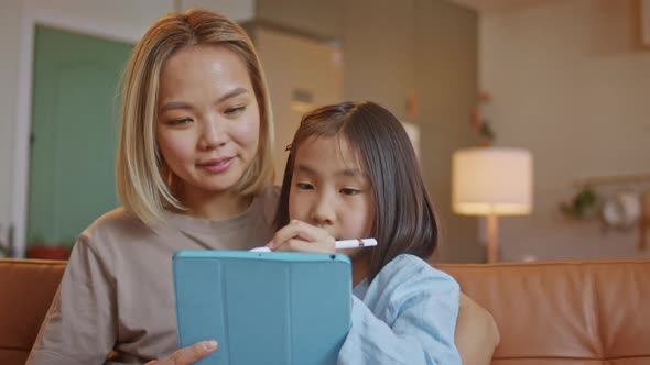 Happy Young Asian Mom and Daughter Doing Homework By Using Tablet Device Draw With Pencel Sitting on