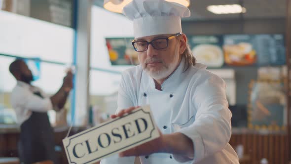 Portrait of Mature Chef in Uniform Turning Closed Sign and Turning Back Open Sign in Modern Cafe