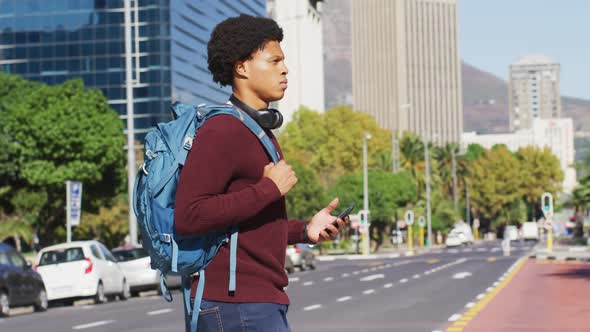 African american man in city, using smartphone, wearing headphones and backpack crossing street