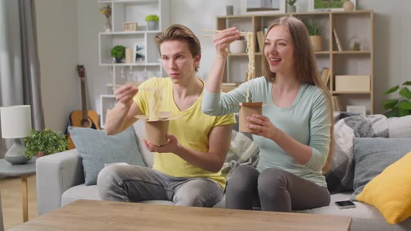 Young Couple Eating Noodles From Box with Chopsticks at Home in the Living Room