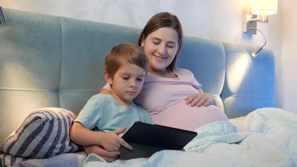 Happy Smiling Little Boy with Pregnant Mother Watching Cartoons on Tablet Computer in Bed at Night