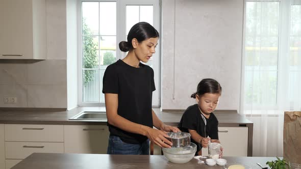 Mom and Daughter are Sifting Flour Through a Sieve Into a Bowl in the Kitchen