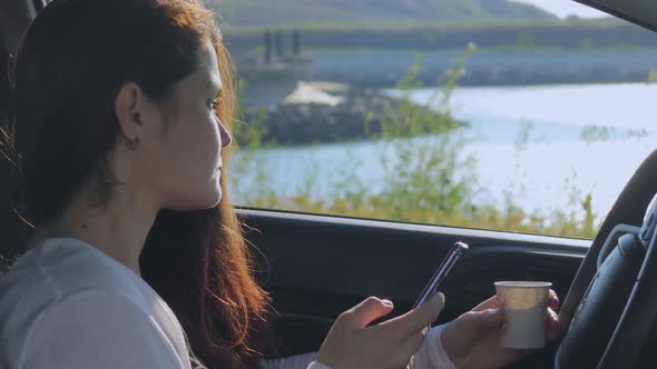 A Young Woman Sits In A Car By The Seashore Drinks Coffee And Uses The Smartphone