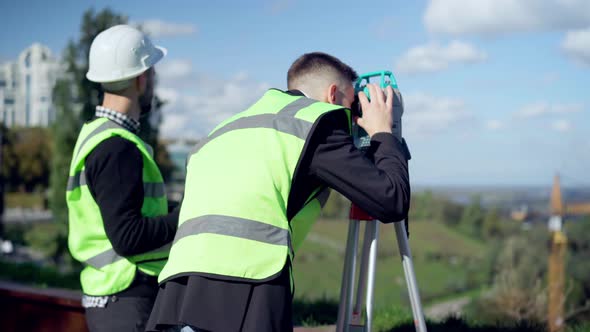 Confident Architect Looking Away with Theodolite As Blurred Engineer with Binoculars Standing at