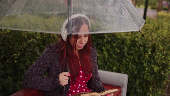 Young Beautiful Woman in Headphones with Transparent Umbrella Reading Book Sitting on Bench in City