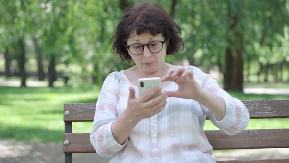 Senior Woman with Poor Eyesight Looking at Smartphone Screen in Sunny Park. Portrait of Female