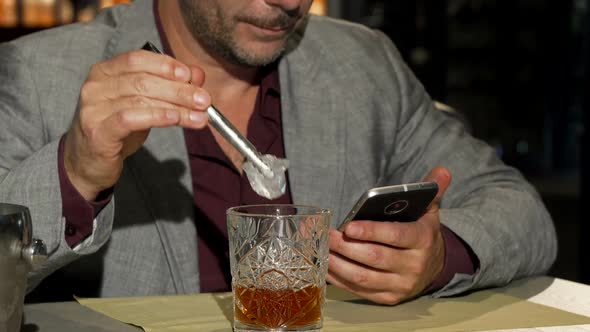 Mature Man Using Smart Phone While Adding Ice Cubes To His Whiskey