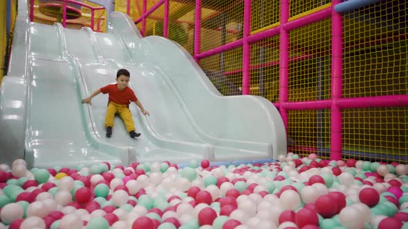 Little Brunette Boy Rolls Down a Slide and Falls Into Balls