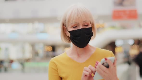 Serious Mature Middle Aged Woman in Medical Mask Walking Holding Smartphone Looking at Screen Typing