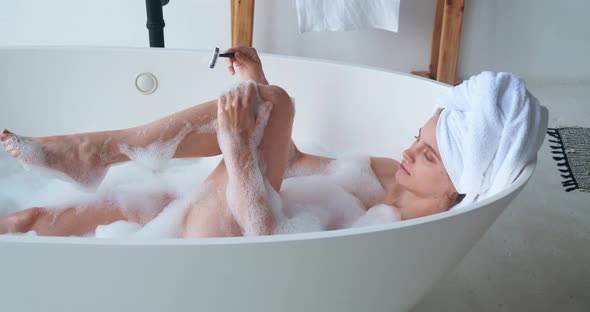 Closeup of Beautiful Woman with Wrapped Head in White Bath Towel Enjoys Foam Bathing Shaving Her Leg