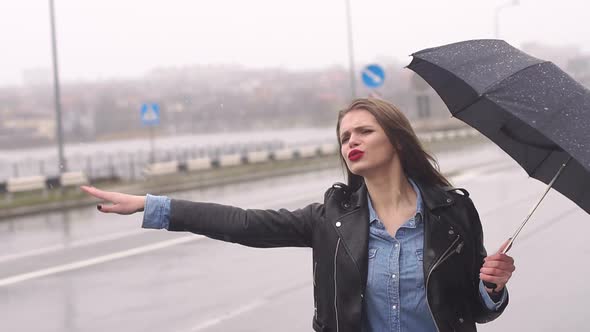 Beautiful Girl on the Road in Heavy Rain Under an Umbrella Trying To Catch a Car