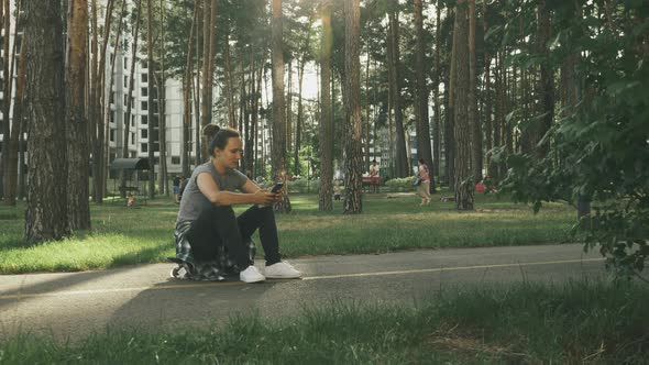 Brunette female sitting on skateboard with phone in hands in city park at sunset.