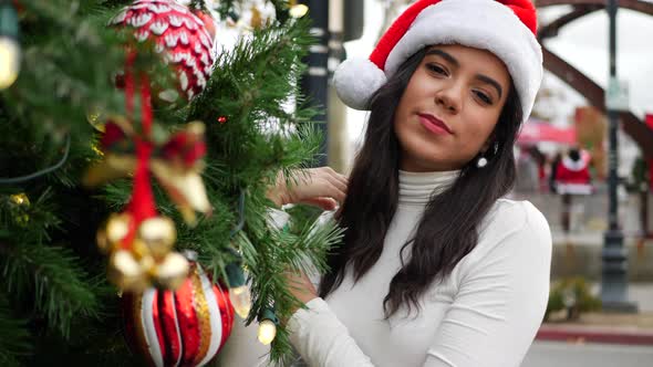 A merry woman in a festive Santa Claus hat decorating a holiday Christmas tree with ornaments, jingl