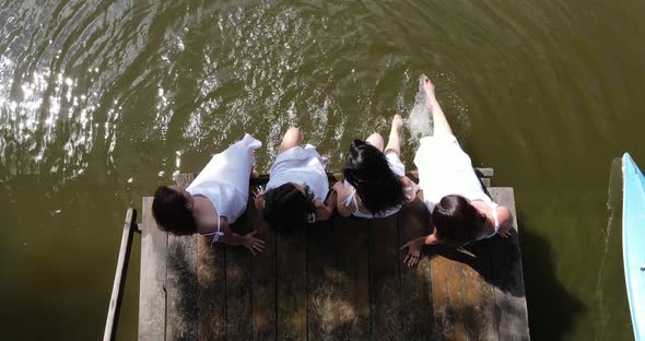 Girls In White Dresses Sit On The Pier And Splash In The Water
