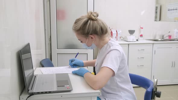 Young Blonde Nurse Wearing Medical Gloves and Mask Writes Down Information About Patient in Hospital