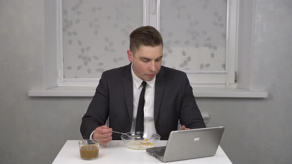 A Young Man in a Suit Eats Cereal and Drinks Coffee and Uses a Laptop